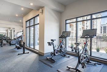 A gym with exercise bikes and a view of a building outside the window at The Delaney at East Park Apartments, Kennesaw, Georgia